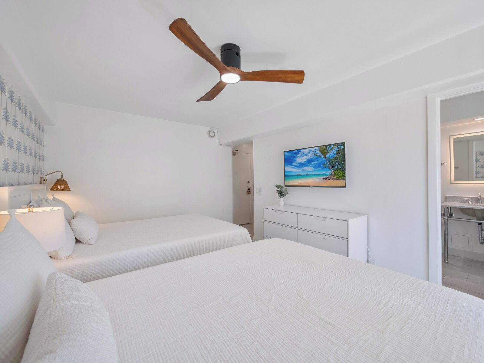 Quarto com duas camas, um ventilador de teto, uma televisão e um banheiro visível por uma porta aberta.