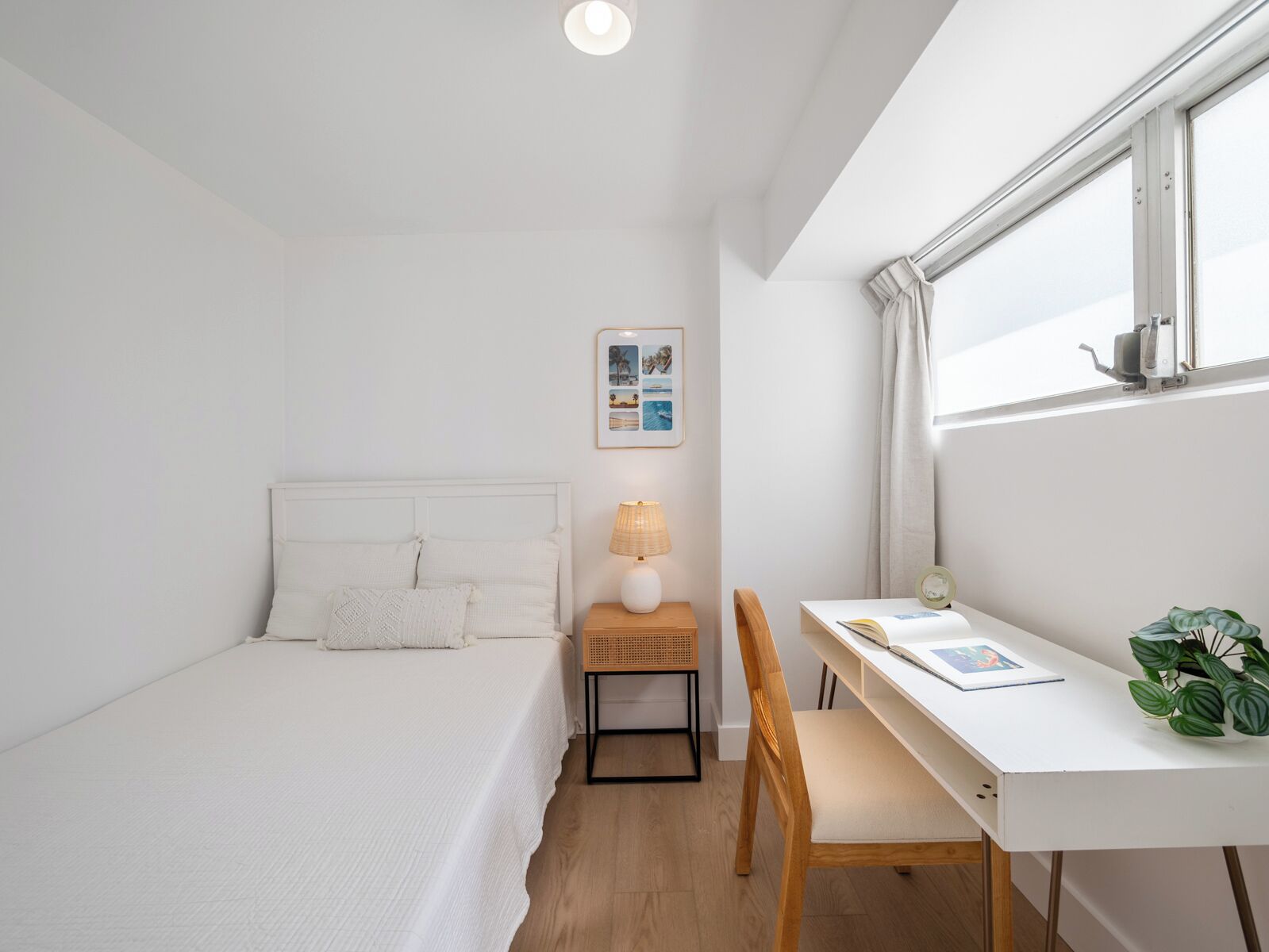 A small, white room with a bed, a desk, a chair, a lamp, a picture frame, and a window.