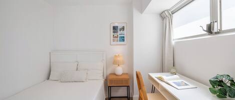 A small, white room with a bed, a desk, a chair, a lamp, a picture frame, and a window.