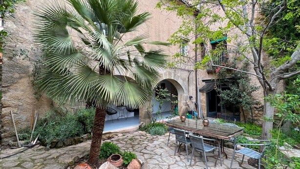 Un patio con una palmera, suelo de piedra, una mesa con sillas y un edificio rústico con arcos.