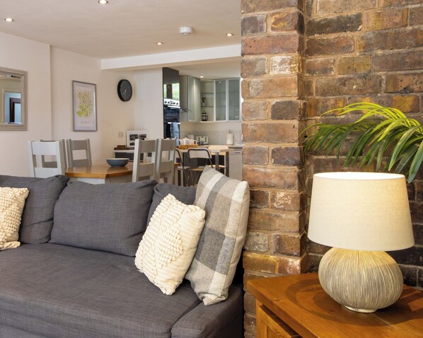 A modern living room with a grey sofa, a wooden side table with a lamp, a brick wall, and a view into the kitchen area. - Jackfield Old School (Jackfield)