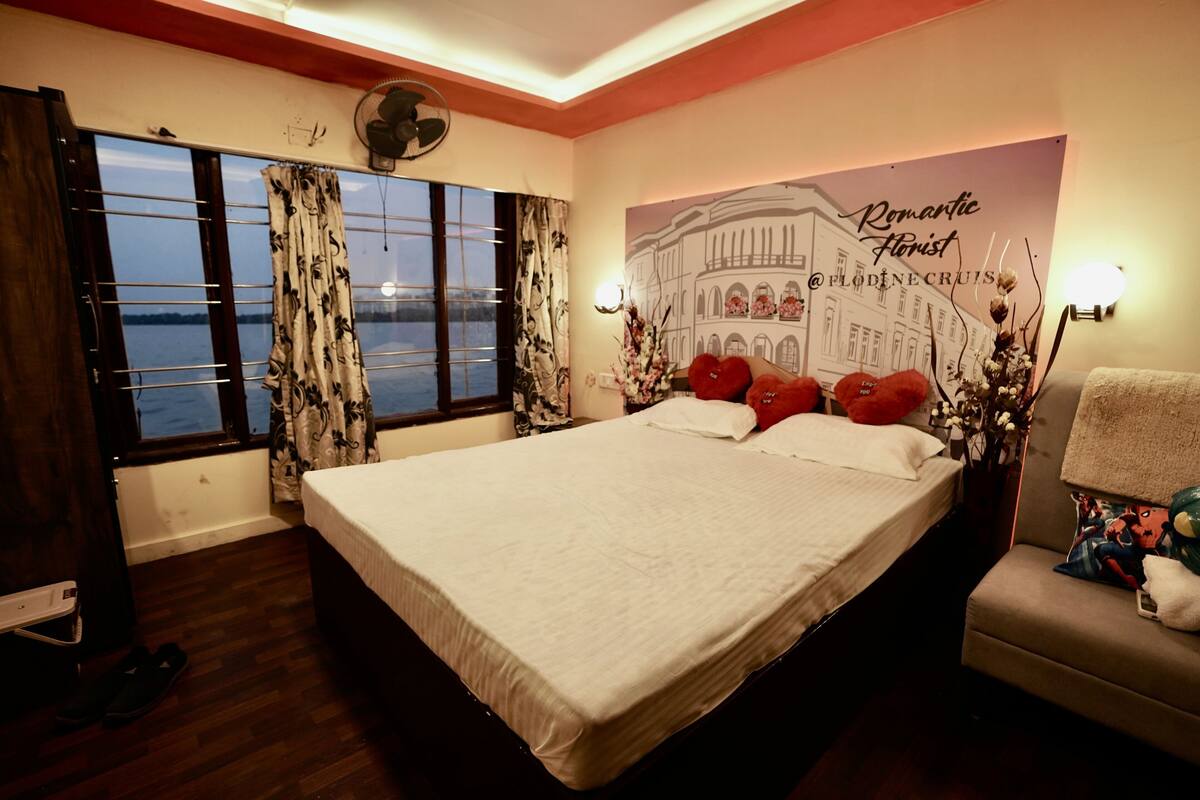 A bedroom with a large bed, a nightstand, a chair, a window with curtains, a wall-mounted fan, and a decorative wall piece.