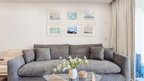 Un sofá gris con una mesa de centro de vidrio, un jarrón con flores y cuadros en la pared.