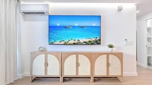 Ein Zimmer mit einem großen Flachbildfernseher an der Wand, der eine tropische Strandansicht zeigt, einem hellen Holzschrank mit Glastüren im Vordergrund und einer weißen Klimaanlage an der Wand.