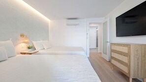 A modern bedroom with two beds, a bedside table with a lamp, a TV on the wall, and a wooden dresser.