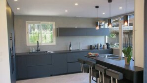 Une cuisine moderne avec des meubles de couleur sombre, un îlot central doté de tabourets, une grande fenêtre et des luminaires suspendus.