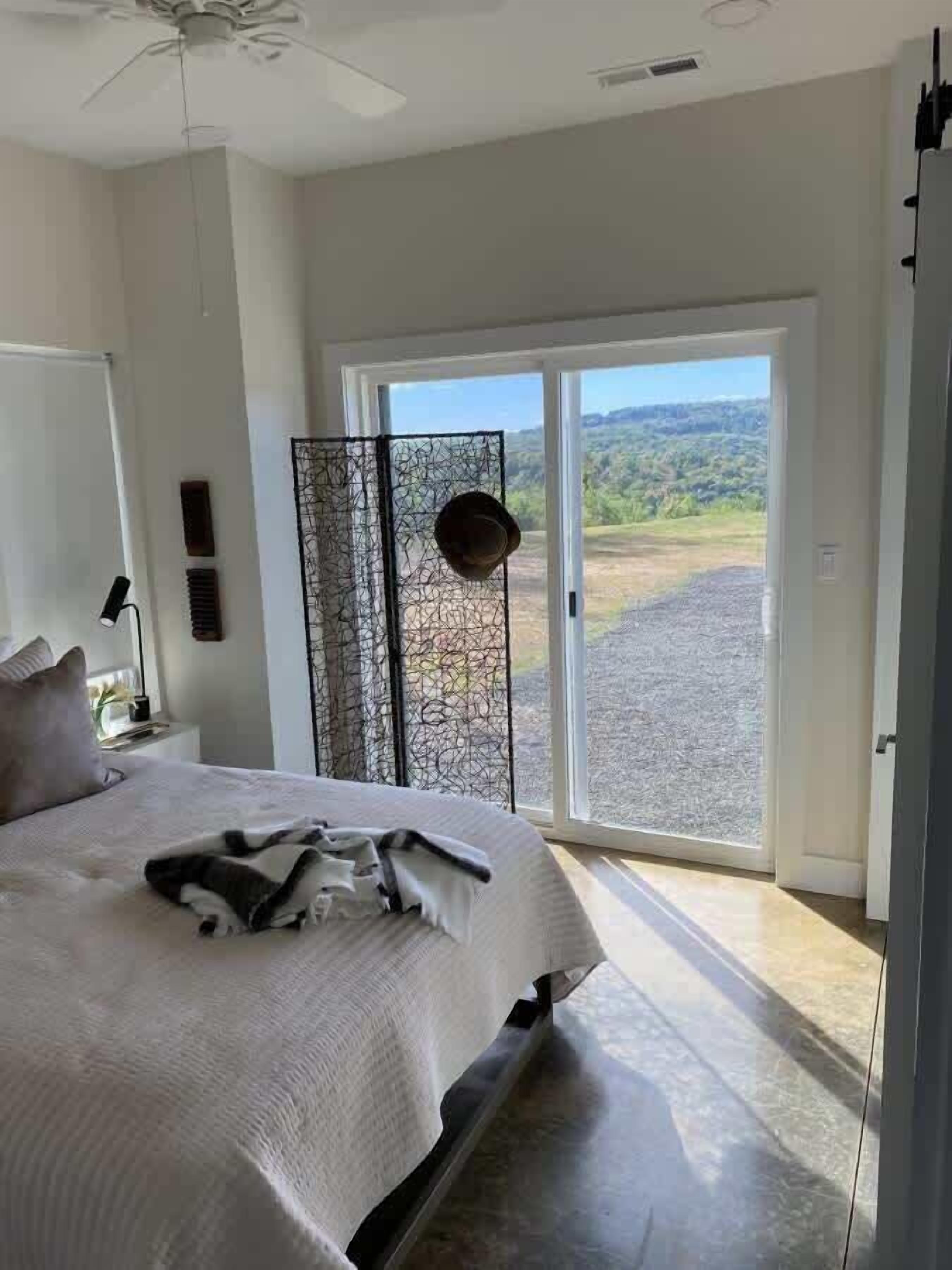 A bedroom with a bed, a ceiling fan, a nightstand, a lamp, a window with a view of the outdoors, and a sliding glass door.
