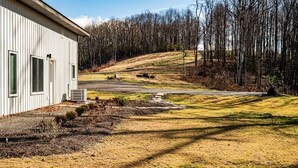 A single-story building with a flat roof, a paved driveway, and a grassy area with a few trees. - The Gathering Barn by Buffalo Mountain Getaway (Meadows of Dan)