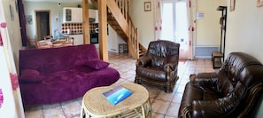 A living room with a purple sofa, a brown leather armchair, and a wicker table. A staircase leads to a kitchen area in the background.