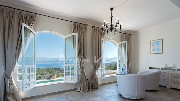 A spacious room with large windows offering a scenic view of the sea and mountains. There is a chandelier, a white sofa, and a small table with decorative items. - Villa with enormous Pool in Kassiopi (Kassiopi)