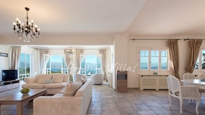 A spacious living room with a sofa, a coffee table, and a TV. There are large windows with curtains, a chandelier, and a dining area with chairs. - Villa with enormous Pool in Kassiopi (Kassiopi)