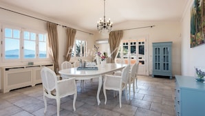 A dining room with a round table, white wicker chairs, a chandelier, and a view of the sea through large windows. - Villa with enormous Pool in Kassiopi (Kassiopi)