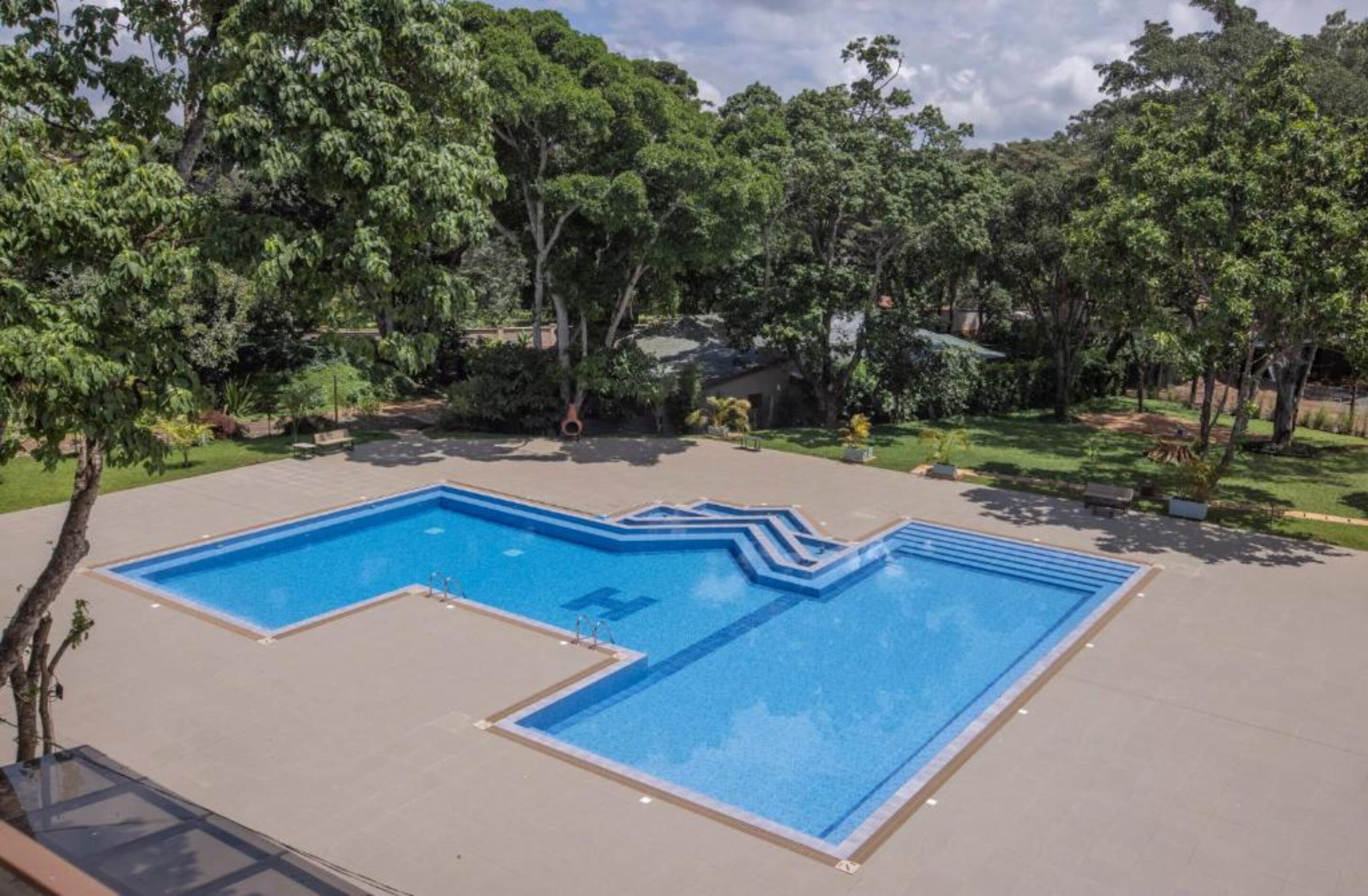 A rectangular swimming pool with a zigzag edge design, surrounded by a paved area with trees and a lawn.
