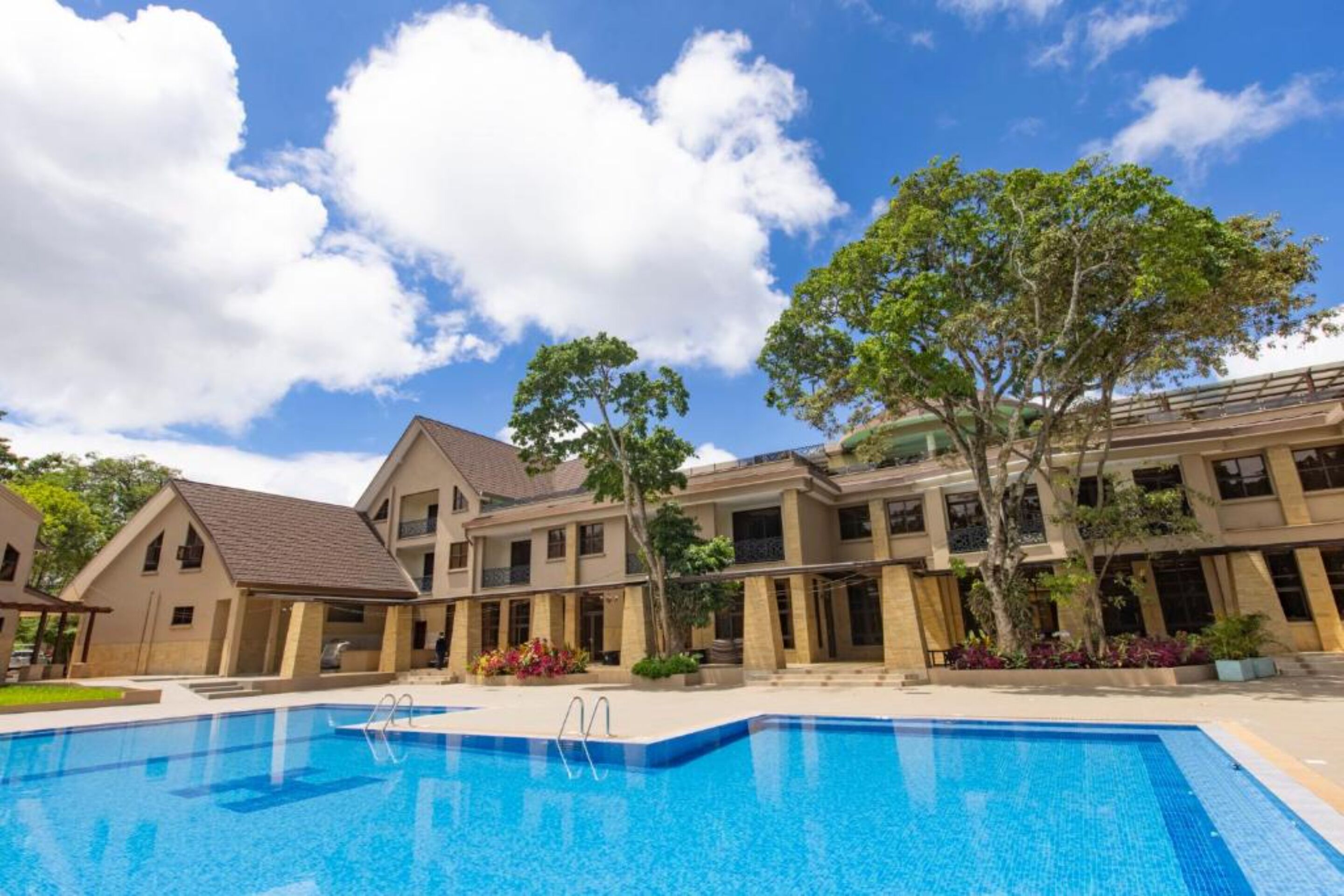A swimming pool with a clear blue water, surrounded by buildings with a beige facade and a large tree in the center.