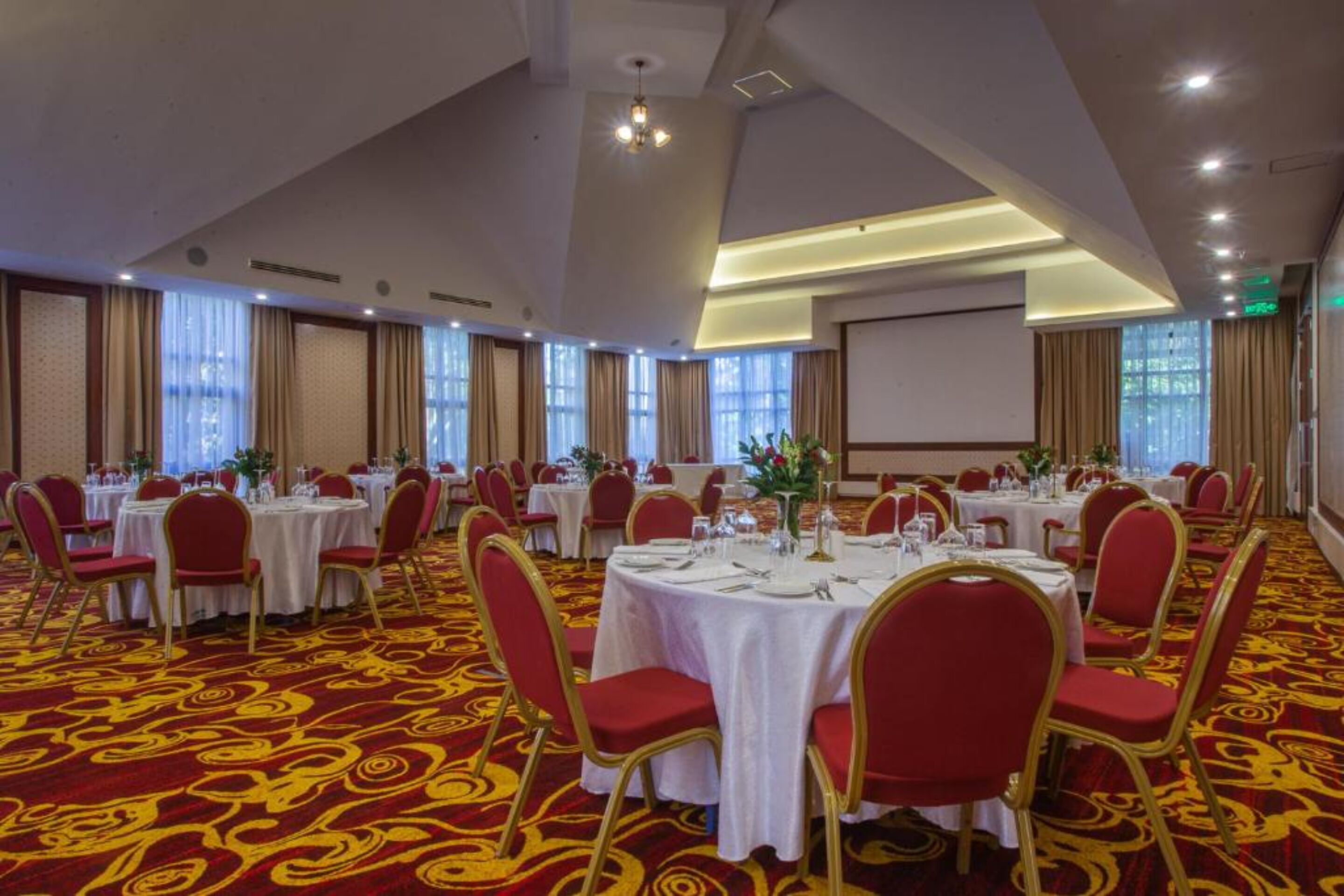 Uma sala de banquetes com mesas redondas, cadeiras vermelhas e piso coberto por um tapete vermelho.