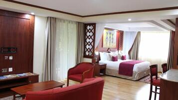 Habitación de hotel con piso de madera, una cama con sábanas blancas y una manta roja, un sillón rojo, un escritorio de madera con una silla y una ventana con cortinas.