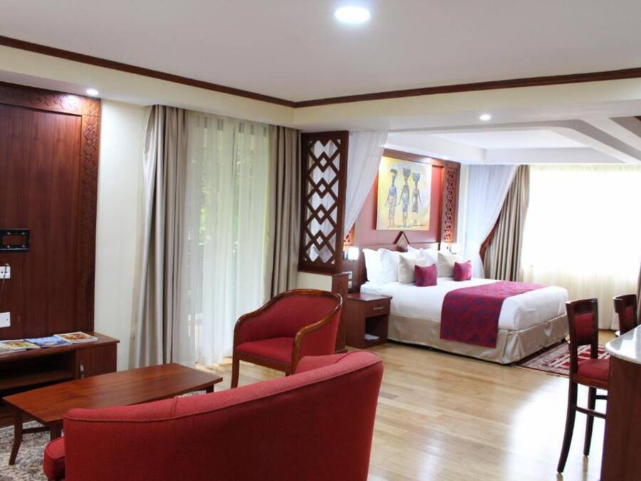 Habitación de hotel con piso de madera, una cama con sábanas blancas y una manta roja, un sillón rojo, un escritorio de madera con una silla y una ventana con cortinas.