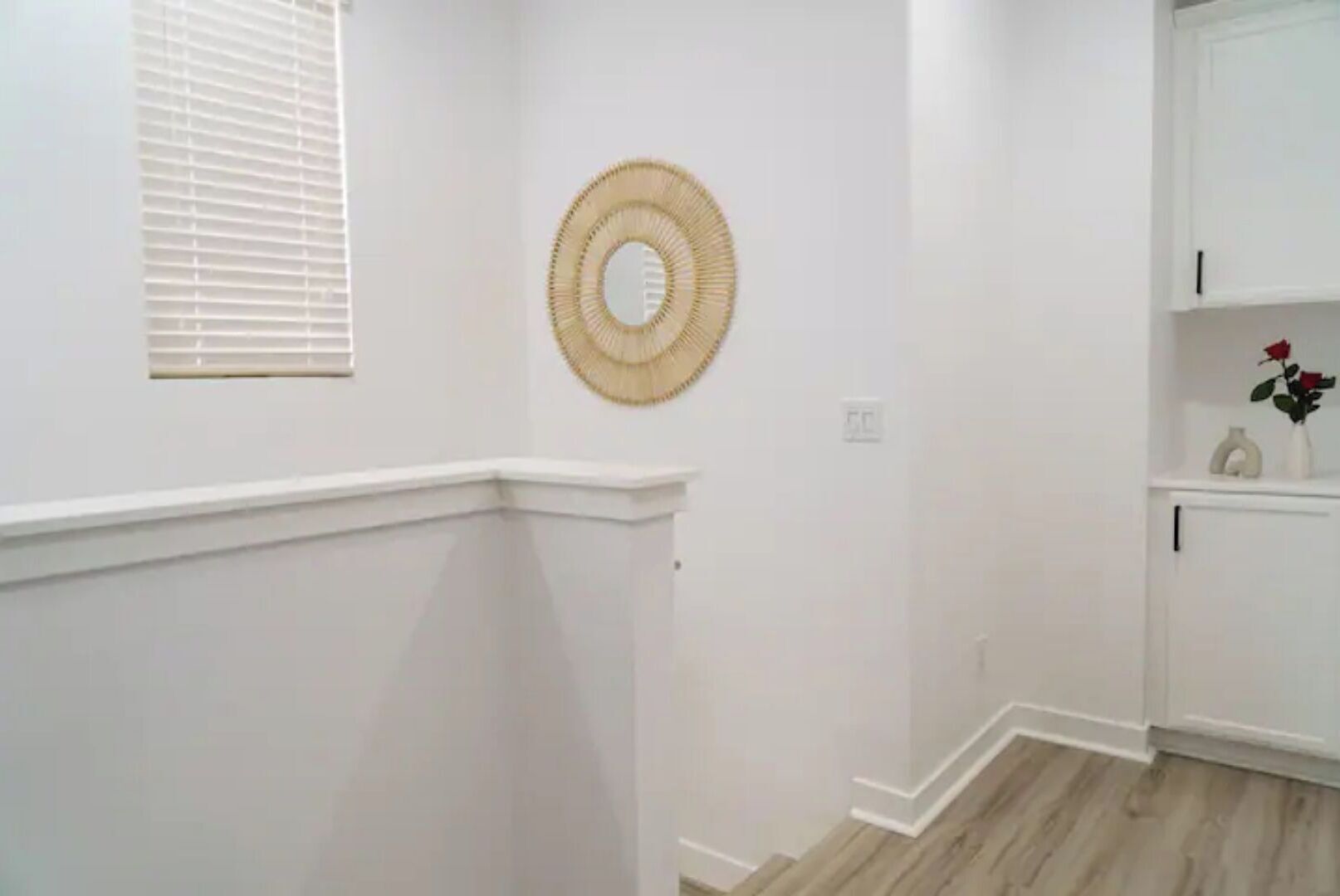 A hallway with white walls, a wooden floor, a white door, and a small shelf with a vase and a rose.