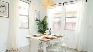 A dining area with a wooden table set for four, surrounded by white chairs, a hanging light fixture, and a potted plant.