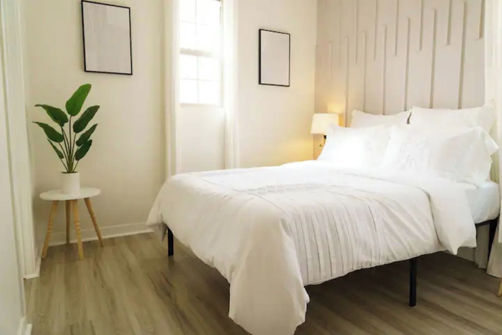 Un lit bien fait, avec une literie blanche, dans une pièce au plancher en bois, agrémentée d’une petite table ronde sur laquelle est posée une plante, et d’une lampe de chevet.