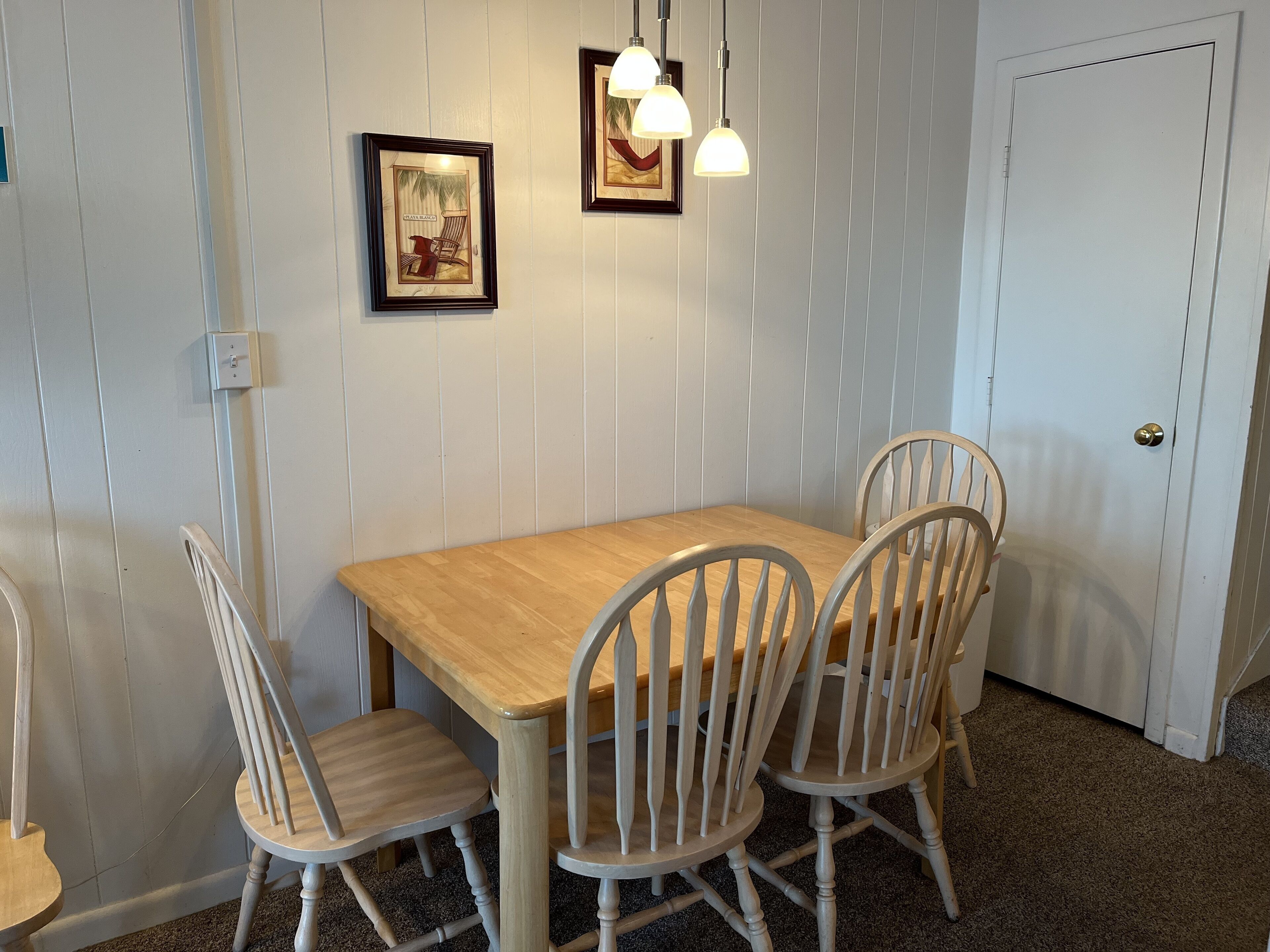 A dining area with a wooden table and four chairs, framed pictures on the wall, and a door in the background.