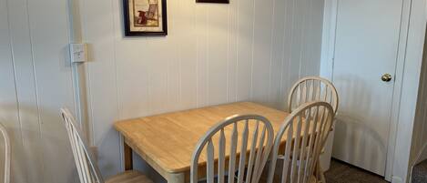 A dining area with a wooden table and four chairs, framed pictures on the wall, and a door in the background.