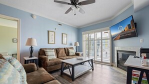 A living room with a brown sofa, a coffee table, a TV mounted on the wall, and a fireplace. - Three bedroom condo with large side balcony allows for viewing of sunrises & sunset (Ocean City)
