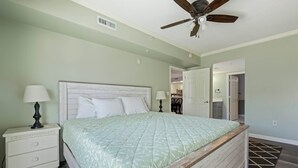 A bedroom with a bed, nightstand, lamp, ceiling fan, and a view of the hallway. - Three bedroom condo with large side balcony allows for viewing of sunrises & sunset (Ocean City)