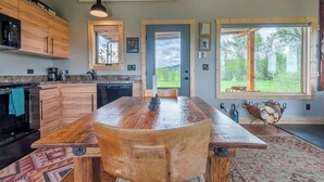A modern kitchen with wooden cabinets, a large island, pendant lighting, and a view of the outdoors through a large window. - Teton Homestead: Ski Cabin w/ Teton Views (Driggs)