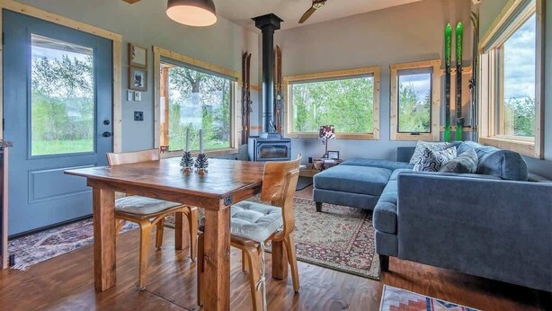 A cozy living room with a wooden dining table, blue sofa, and large windows offering a view of the outdoors. - Teton Homestead: Ski Cabin w/ Teton Views (Driggs)