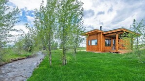 A wooden cabin with a porch, a window, and a chimney, surrounded by a grassy area and trees. - Teton Homestead: Ski Cabin w/ Teton Views (Driggs)