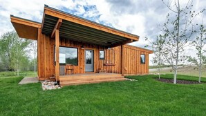 A modern wooden cabin with a covered porch, large windows, and a small outdoor seating area. - Teton Homestead: Ski Cabin w/ Teton Views (Driggs)