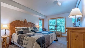 A bedroom with a large bed, wooden headboard, two windows with blinds, a wooden dresser, and a lamp on a bedside table.