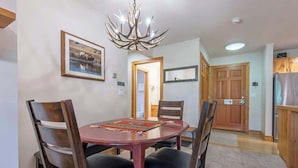 A dining area with a round table, four chairs, a chandelier, a framed picture on the wall, and a kitchen area with wooden cabinets and a refrigerator.