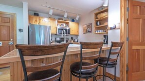A kitchen with wooden cabinets, a stainless steel refrigerator, a microwave, and a sink. There are three wooden bar stools with black metal frames.