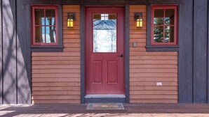 A wooden house with a red door, two windows with red frames, and two wall-mounted lanterns. - Teton Homestead: Bunkbeds, Private Deck, Targhee (Driggs)