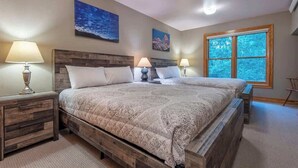 A bedroom with a wooden bed frame, two bedside tables with lamps, and a large window with a view of trees. - Teton Homestead: Modern Corner Top Unit + Hot Tub (Driggs)