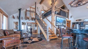A spacious living area with a large fireplace, a wooden staircase, and a bar area. - Teton Homestead: Western Cabin w/ Hot Tub & Views (Driggs)