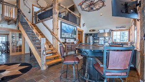 A spacious living area with a large wooden staircase, a bar area with stools, and a kitchen in the background. - Teton Homestead: Western Cabin w/ Hot Tub & Views (Driggs)