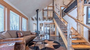 A spacious living room with a large window, a wooden staircase, and a stone fireplace. - Teton Homestead: Western Cabin w/ Hot Tub & Views (Driggs)