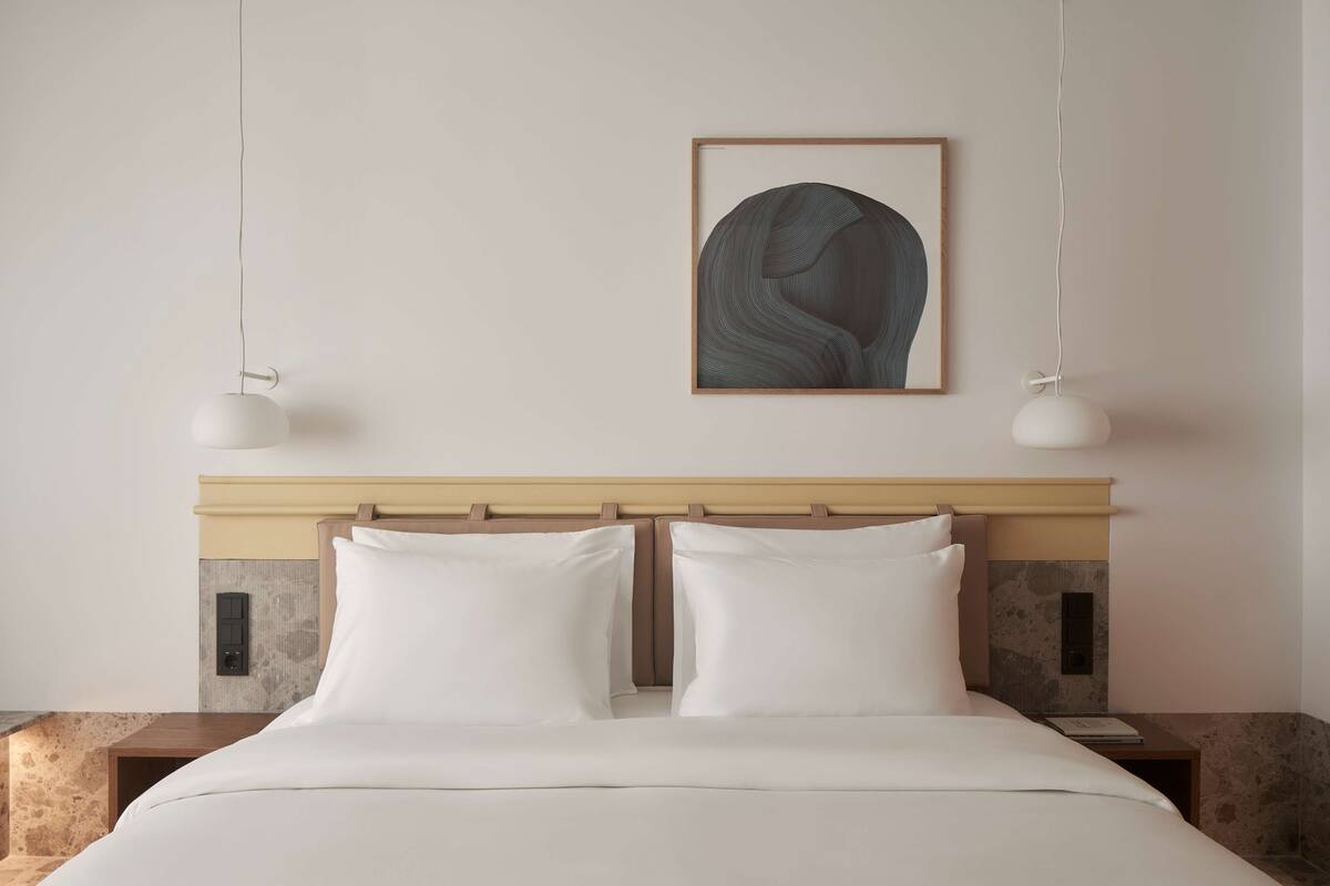 A neatly made bed with white linens, a wooden headboard, and a framed abstract painting above.
