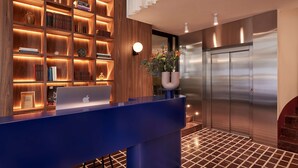 A reception area with a blue counter, wooden shelving with books and decorative items, a potted plant, and a modern elevator. - The Social Athens Hotel, a member of Radisson Individuals (Athens)