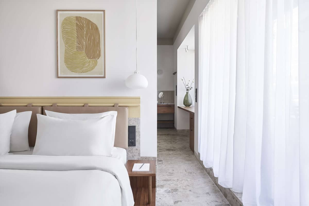 A hotel room with a bed, a bedside table, a framed artwork, and a view of a bathroom and kitchen area.