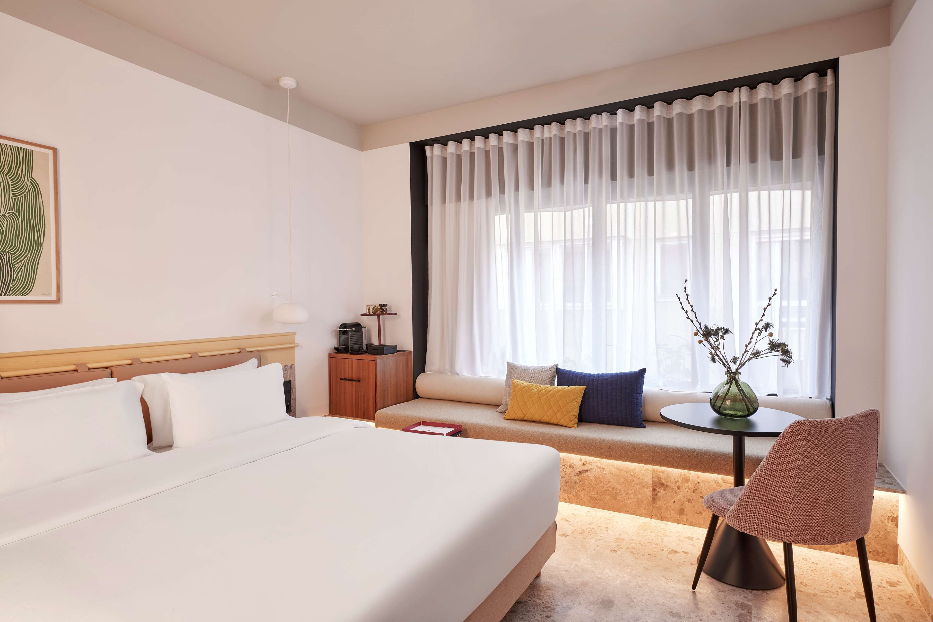 A hotel room with a large bed, a sofa, a small table, and a chair. There is a window with sheer curtains and a vase with flowers on the table.