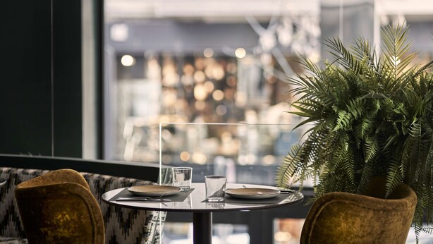 A dining area with a round table set for two, a potted plant, and a view of the city through large windows. - The Social Athens Hotel, a member of Radisson Individuals (Athens)