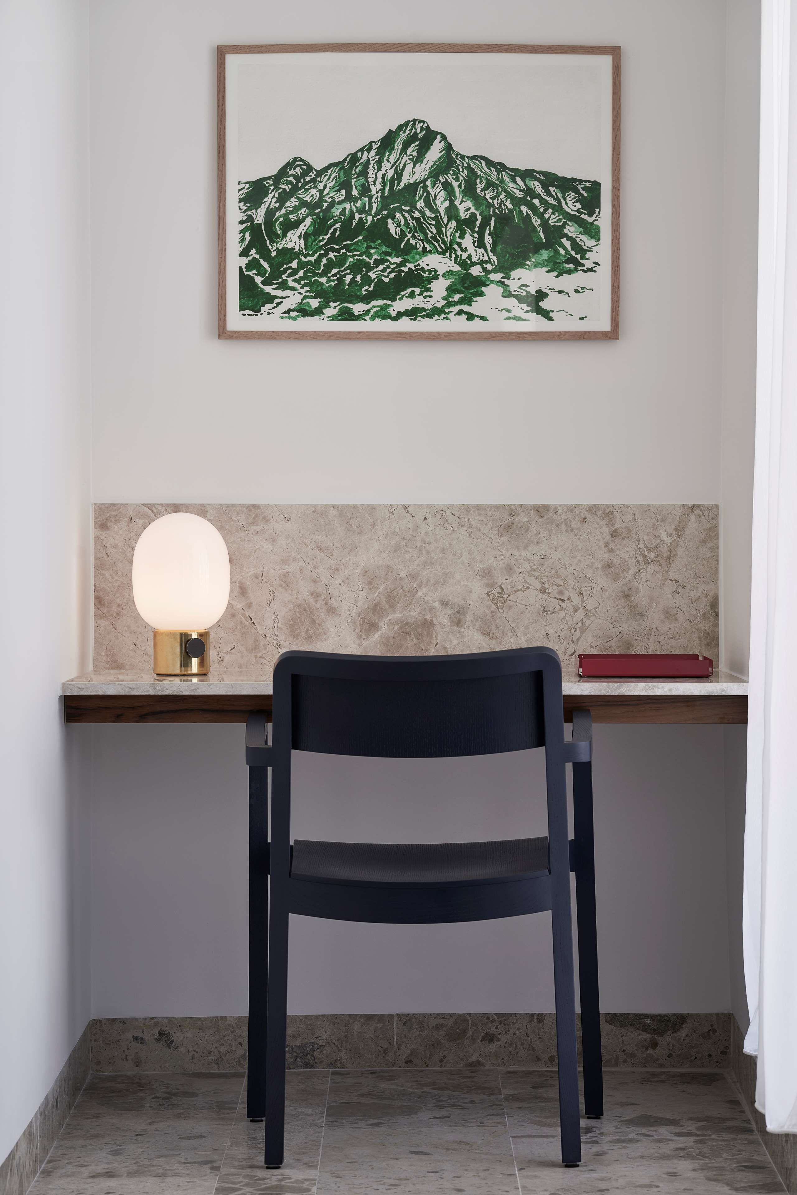 A desk with a chair, a framed mountain artwork, a round table lamp, and a red book.