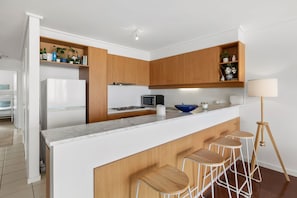 A modern kitchen with wooden cabinets, a white countertop, a built-in microwave, and a blue bowl. - Apartment 30 Pacific Apartments (Lorne)
