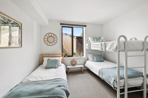 A room with two single beds, a bunk bed, a window with blinds, a small table with a plant, and a wall-mounted decorative item. - Apartment 30 Pacific Apartments (Lorne)