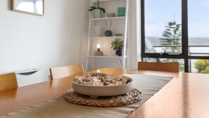 A dining table with a bowl of seashells on a woven placemat, a white bookshelf with decorative items, and a view of the sea through a large window. - Apartment 30 Pacific Apartments (Lorne)
