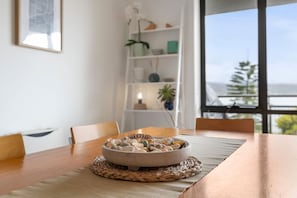 A dining table with a bowl of seashells on a woven placemat, a white bookshelf with decorative items, and a view of the sea through a large window. - Apartment 30 Pacific Apartments (Lorne)